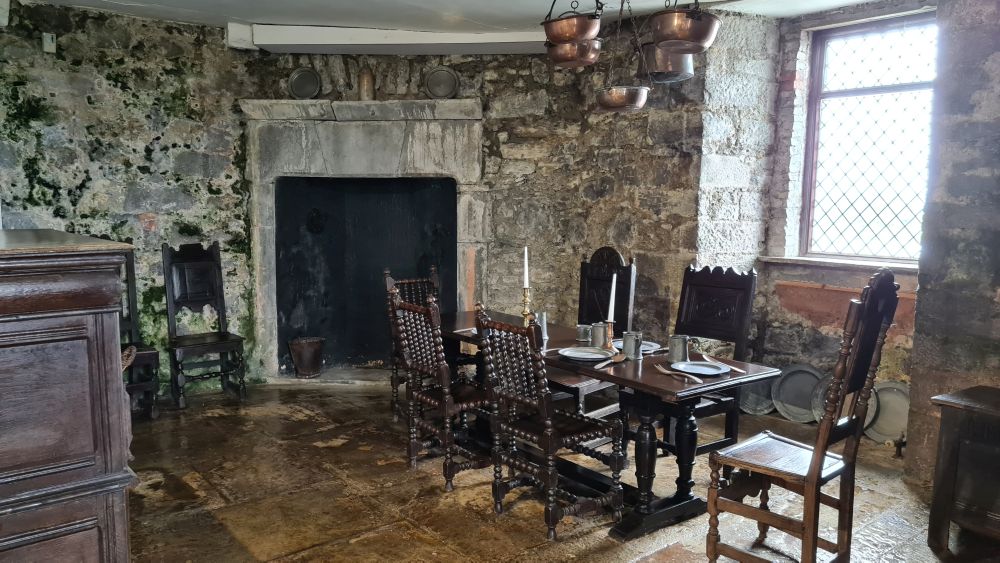 A table with heavy oak chairs around it, copper pots hanging from the ceiling, a fireplace.