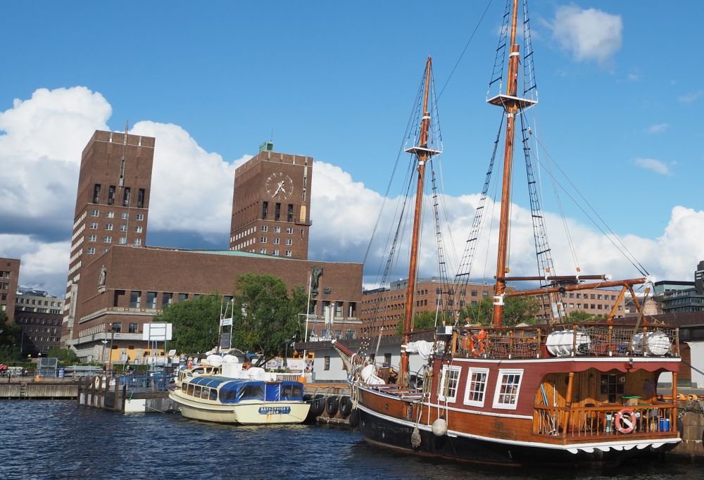 Square brick building with two square towers, seen across the harbor, where several boats are docked.