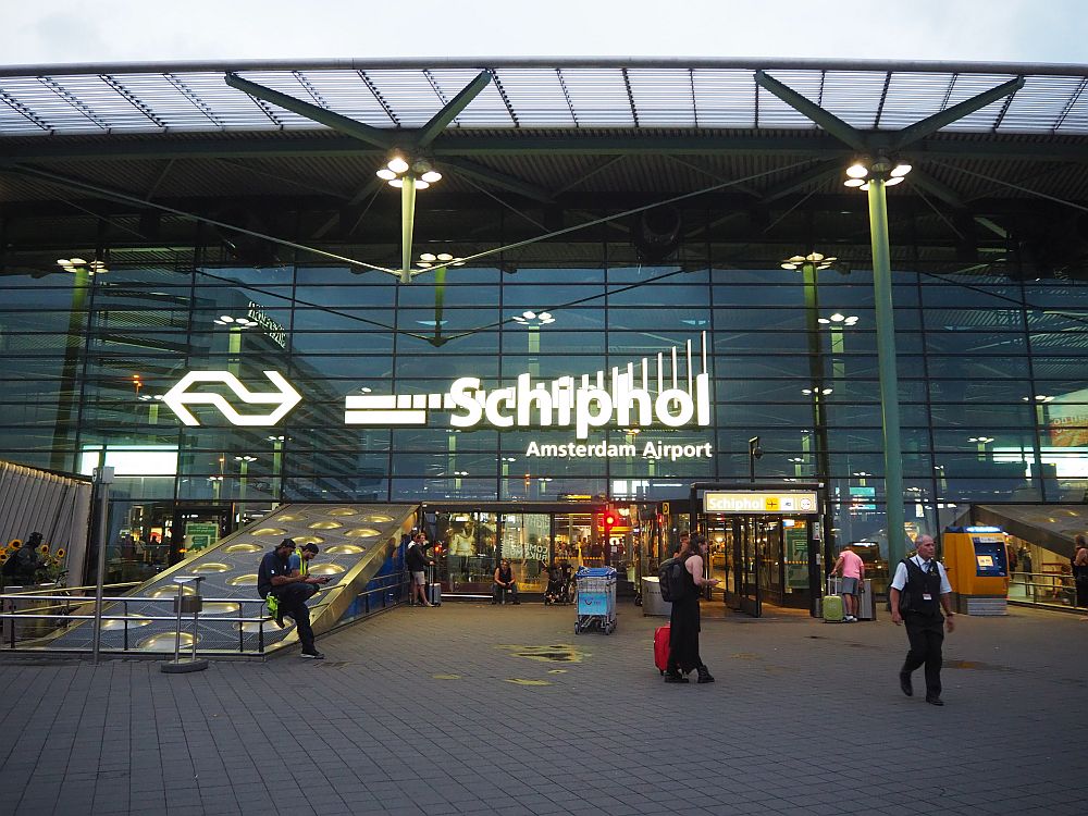 Shiny dark glass building with the word Schiphol on it. People going in and out.