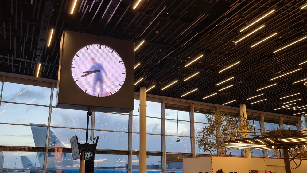 A large clock hanging from the ceiling, with a white face and visible through the clockface, a man dressed in coveralls who is painting one of the hands of the clock.