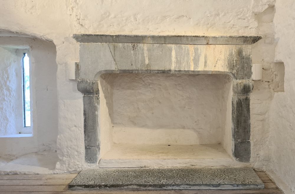 A gray stone fireplace set in a white-plastered wall. A tall narrow window next to it.