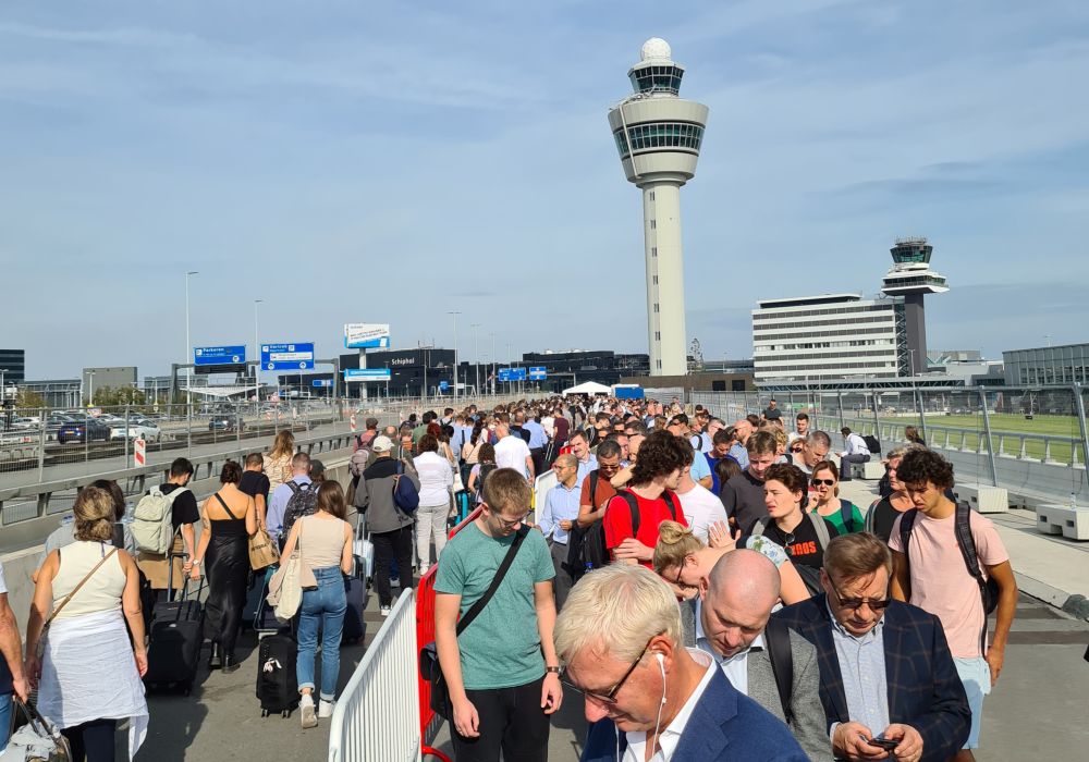 Many people in line outside, some coming toward the camera, some away. In the distance, the control tower with the airport behind it.