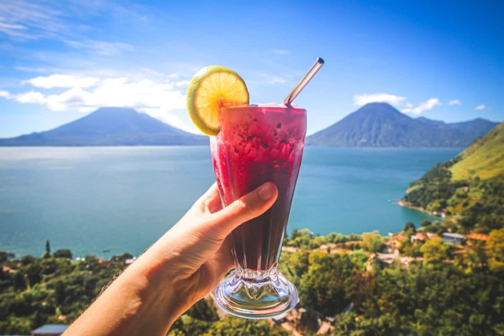 A hand holding a red drink up in front of sea and mountains scenery.