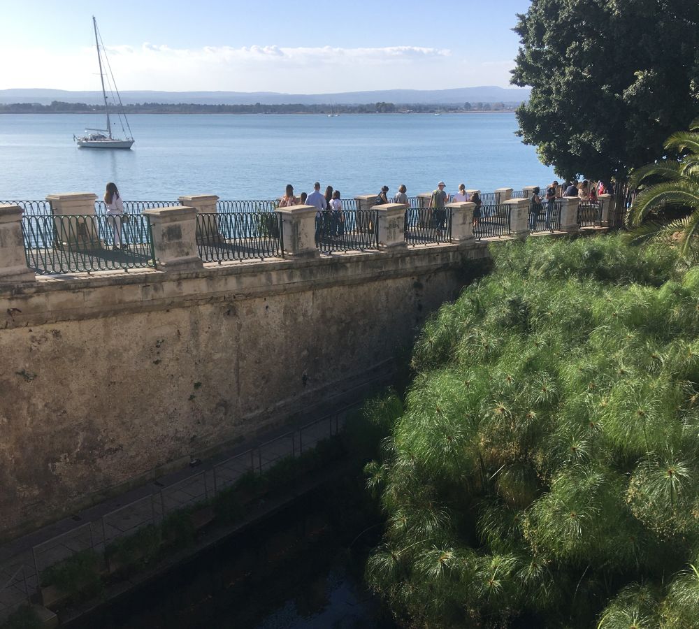 Water below, trees to the right, a high wall with people walking on it behind. Sea and a boat beyond the wall.