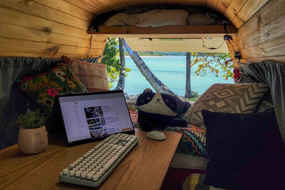 Inside a camper, a desk with a laptop open on it. Beyond, the back of the camper is open on a view of the ocean.