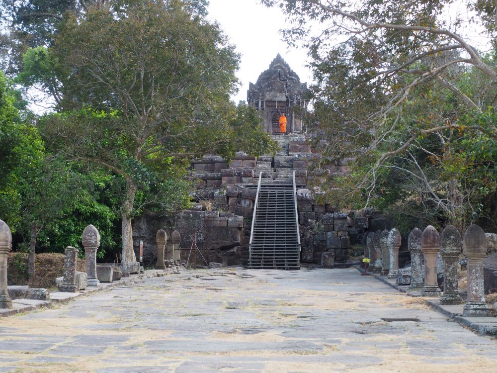 A wide walkway with standing stones along the sides, and a temple on a hill straight ahead, with a monk in orange in front of it.