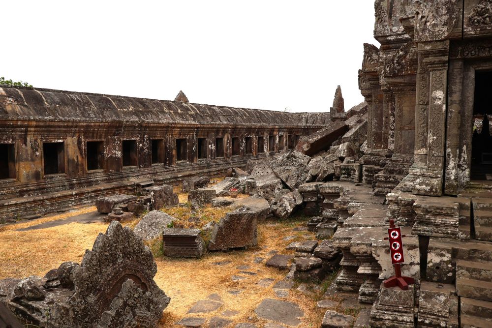 On the left, a stone gallery with roof; on the right, a temple with a pile of large stones next to it.