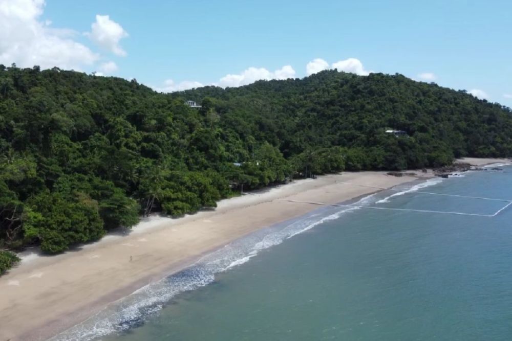 Drone view along a strip of almost-empty white sand edged by the sea and dense rainforest.