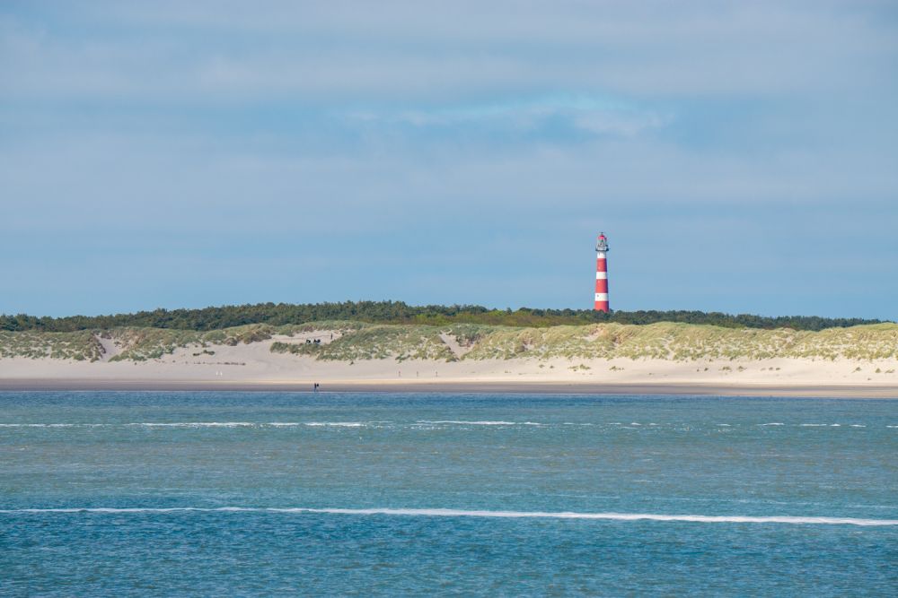 A view from the water of the shore: long white beach, dunes along the beach, and the tall red and white striped lighthouse rising behind the dunes.