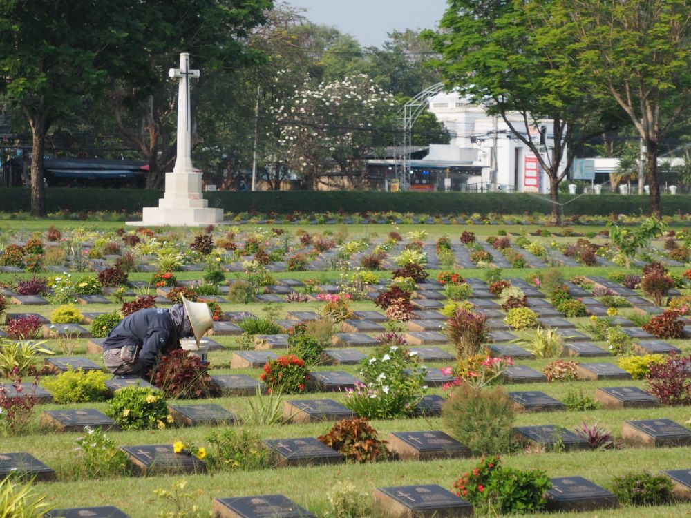 A green flat field with very neat lines of low bronze plaques with flowers between them. A groundskeeper kneels, weeding.