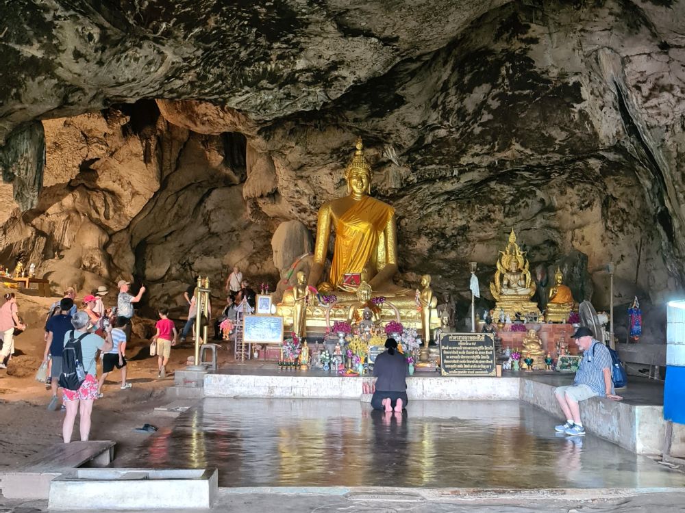 A large sitting Buddha statue with a person kneeling in front of it, praying, and other people, obviously Western tourists, here and there.