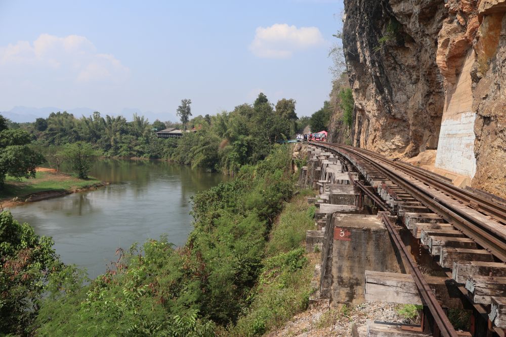 A river down below on the left, a cut rock face on the right, with a wooden structure holding the train tracks along the edge of the rock face.