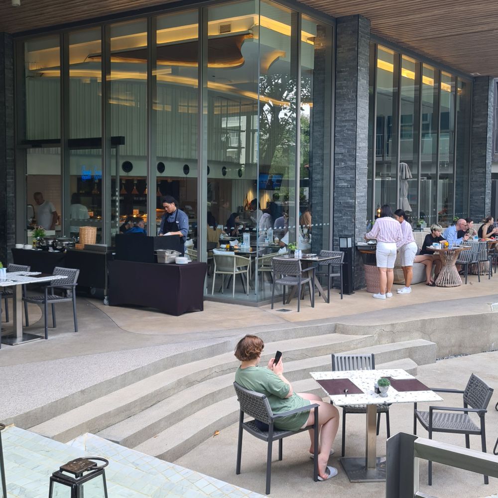 People at scattered tables outdoors, glass walls behind them and people at tables indoors behind the glass.