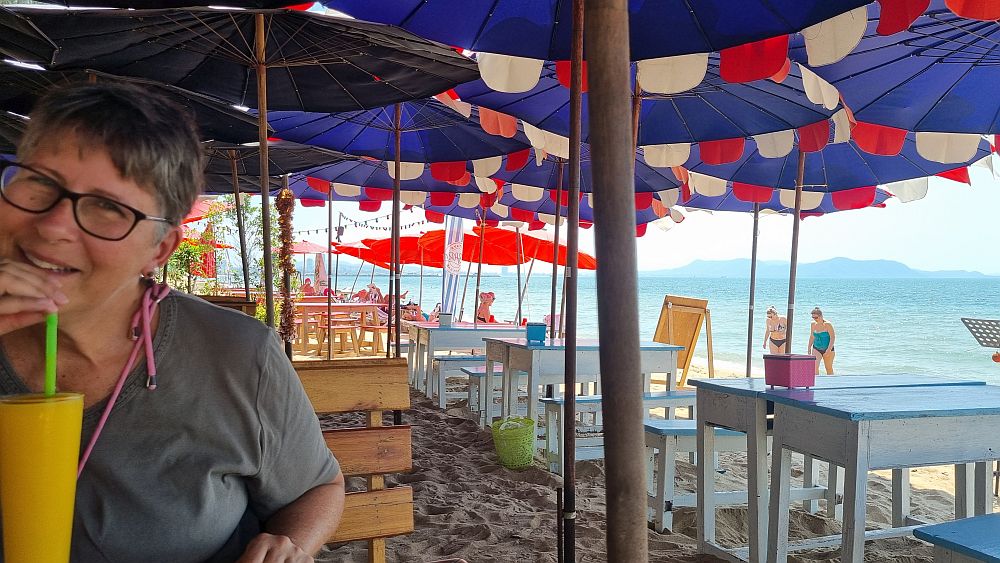 Me drinking a smoothie with, in the background, tables and chairs in sand, shaded by colorful umbrellas.