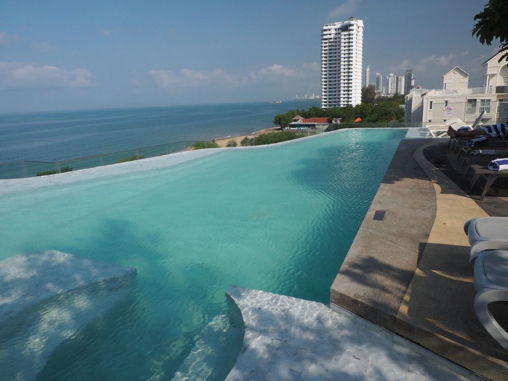 Blue water in an irregularly-shaped infinity pool, with the darker blue of the sea beyond, and a tall apartment building as well.