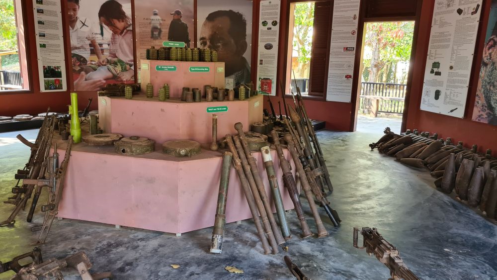 A raised section in the middle of the room has landmines sitting on its shelves and guns leaning against it. The walls behind have text and photos and more landmines on the floor.