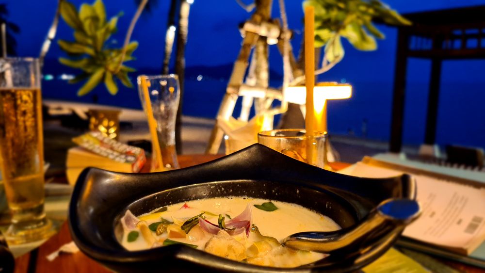 A bowl of soup in the foreground, blurred drinks, lights and dark sky in the background.