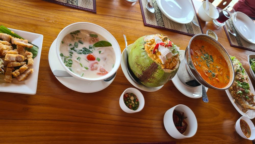 L-R: some crispy pork pieces, a coconut soup, a coconut cut at the top and holding a curry, a bowl of prawns in sauce, and fried fish. 