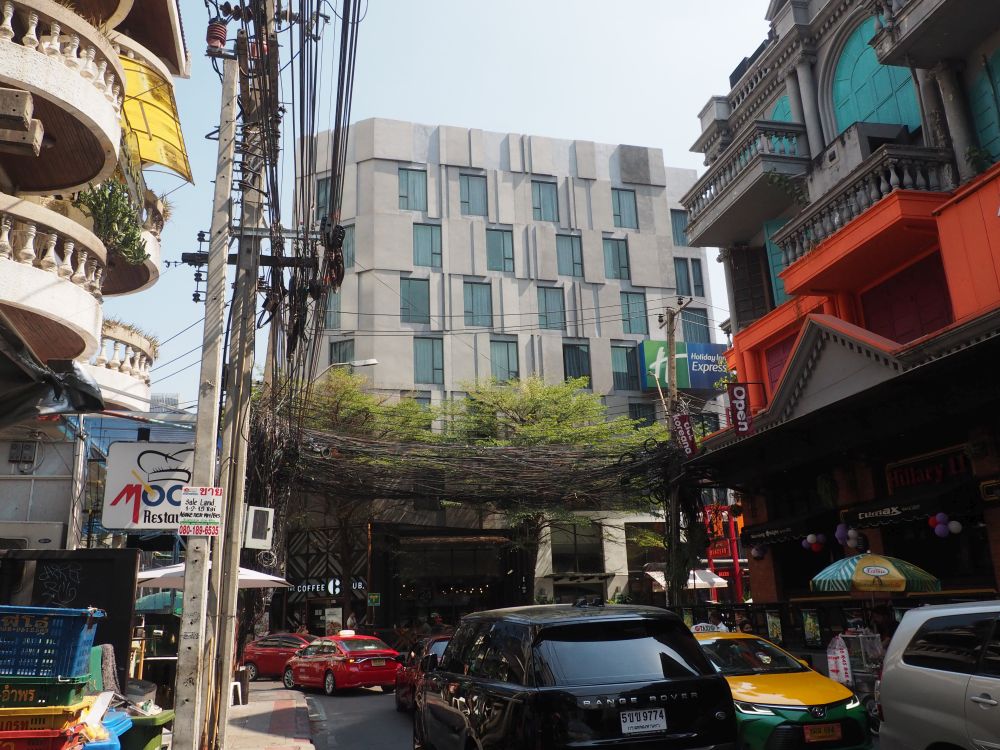 The hotel, about seven storeys, is an odd brutalist-type style. View from a street full of traffic and shops and clubs on either side.