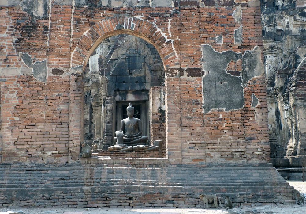 An archway in a brick wall frames a sitting Buddha statue. Several monkeys sit in or nearby the archway. 