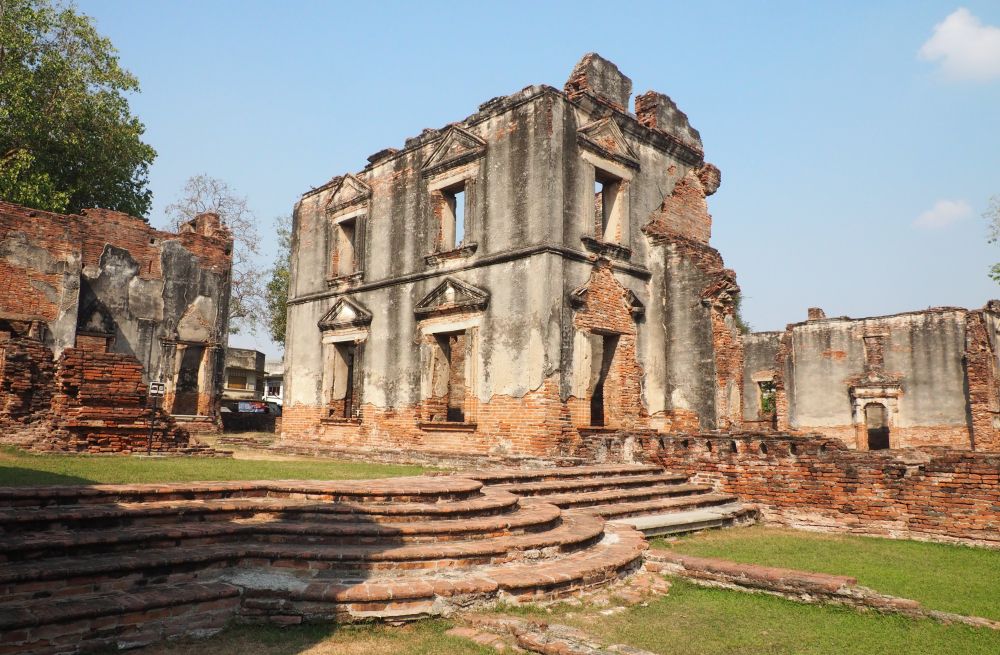 A two-storey structure, brick showing through where the outer cement layer is gone. The windows have decorated tops and the roof is gone.