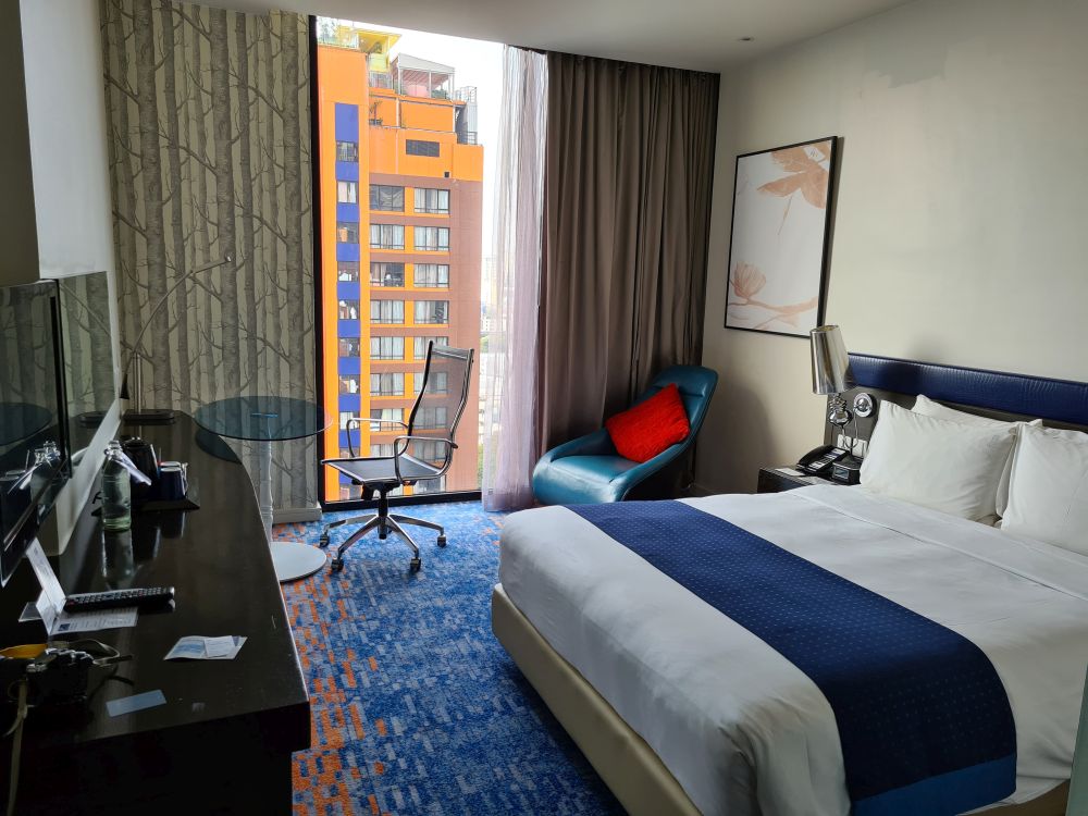 hotel room with large bed, comfy chair in one corner, desk chair in the other, and a floor-to-ceiling window across one wall.