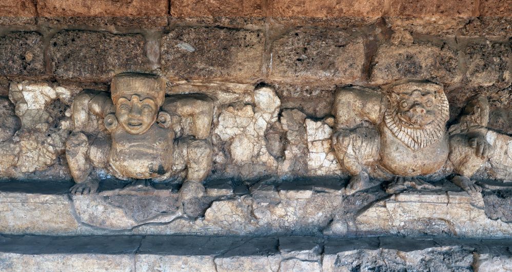 Two stucco figures in a rough stone wall - both squatting. Left has a human face, right has an animal face.