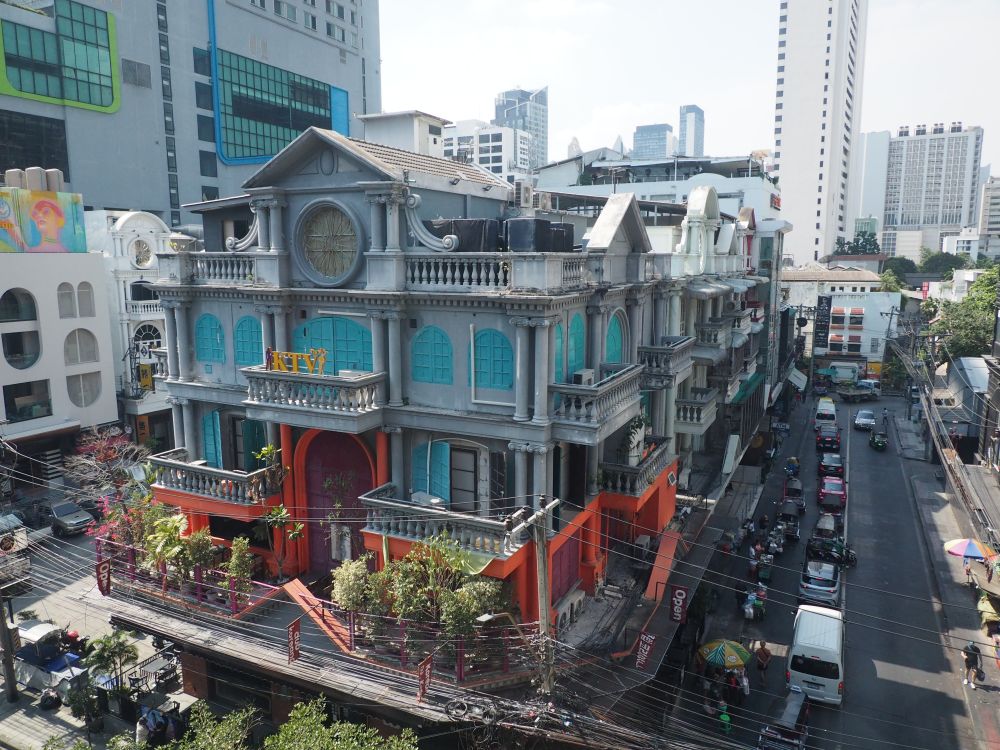 A strange building with lots of potted plants on and around it looks like it once must have been a theater. A street busy with cars.