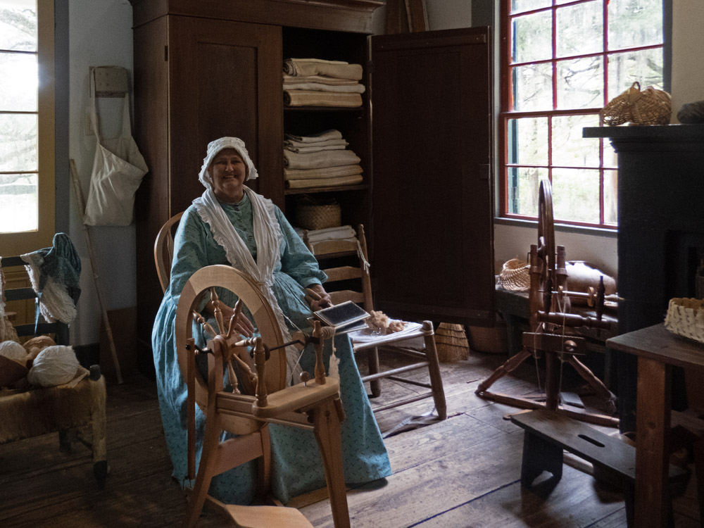 A woman in a long, blue period dress with a bonnet on her head sites next to a spinning wheel, smiling at the camera.