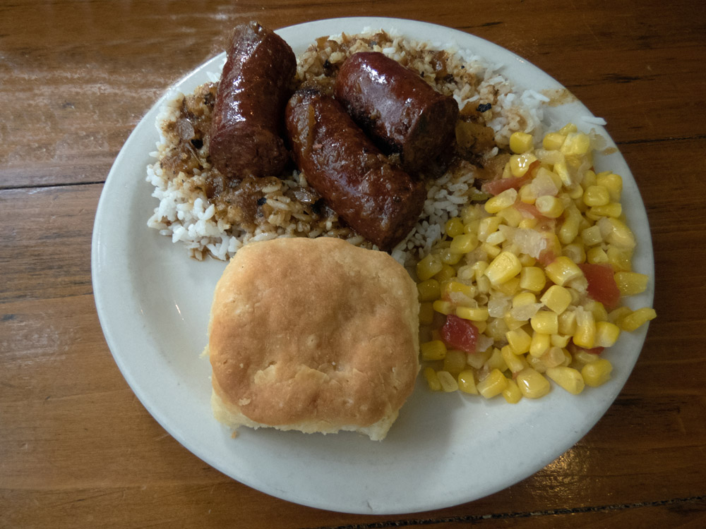 A plate of food: corn, a biscuit, and sausages on rice.