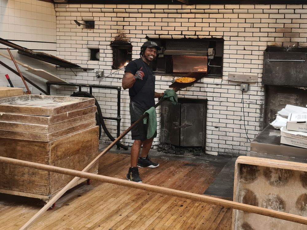 A man stands next to an oven in a wall, holding a long-handled paddle with a pizza on it.