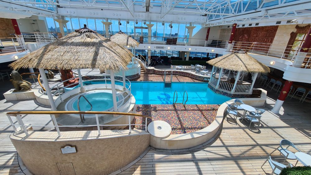 A pool with a hot tub on the near side covered by a thatched roof.