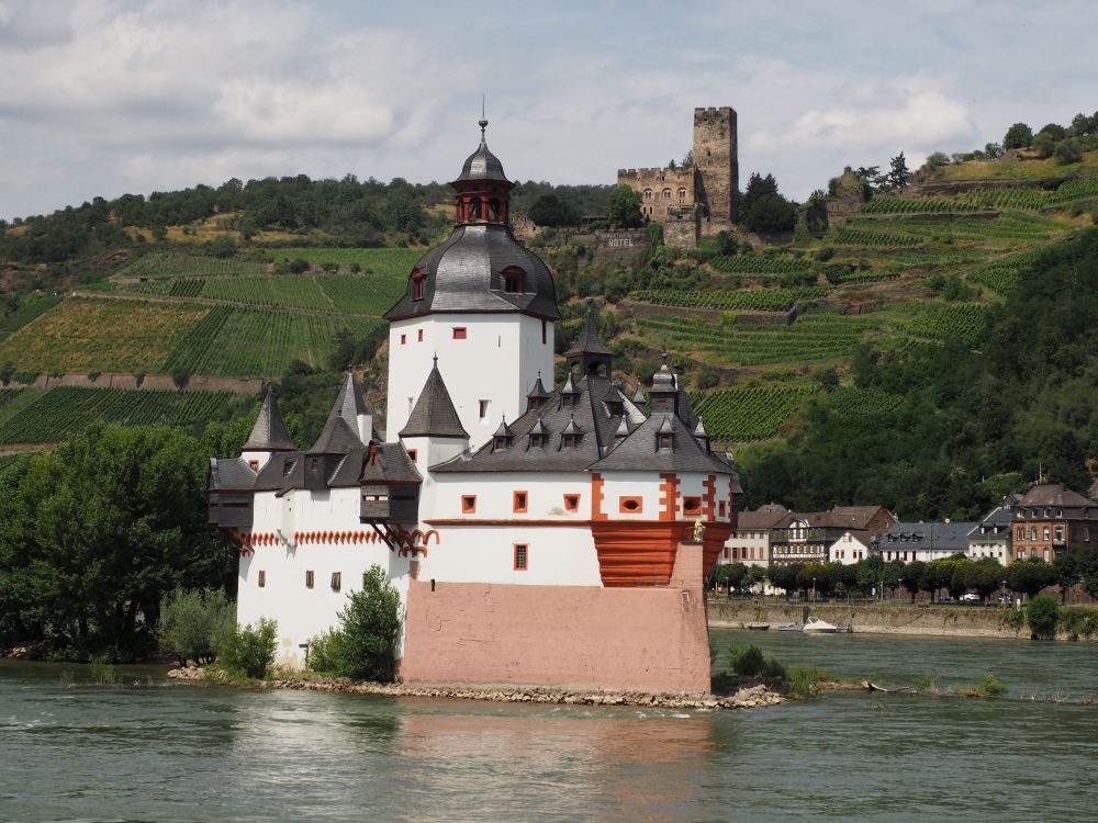 A small white castle at river level, and a medieval stone castle beyond it on a hill.