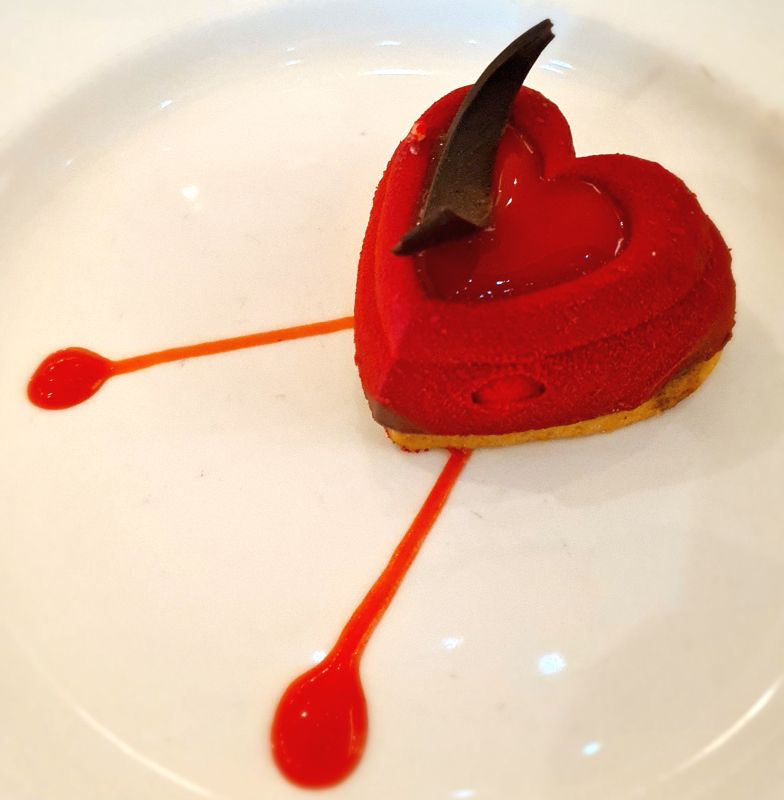 A red heart-shaped pastry, with a piece of chocolate on it and some dribbles of red sauce.