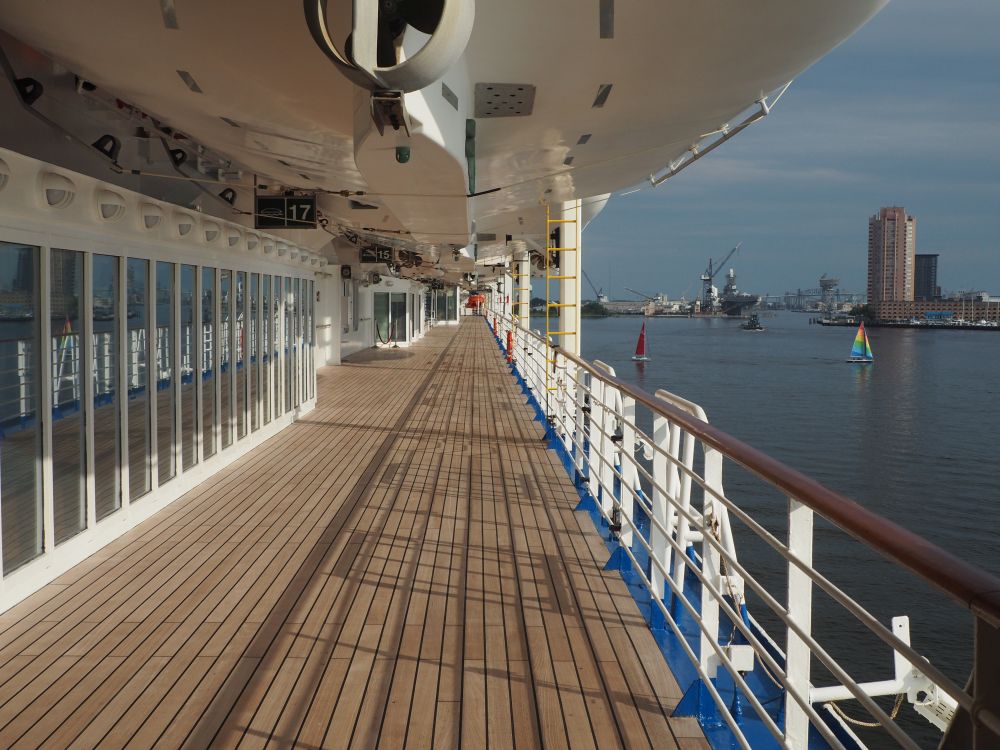 A wide walkway with, on the right, a railing over the water. On the right, windows of the ship. Above, the bottoms of boats.