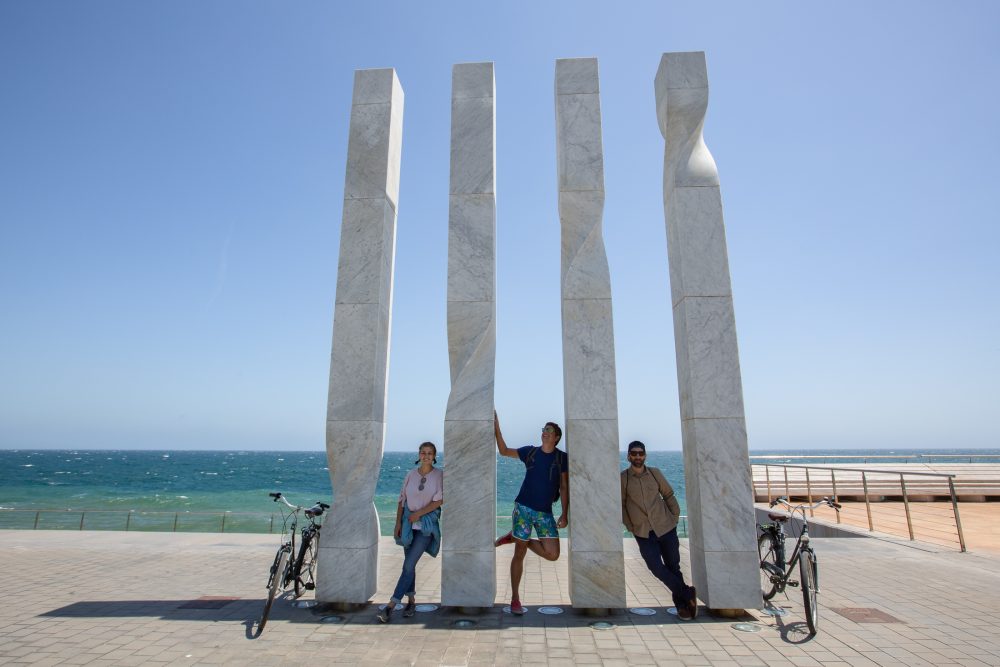 4 white pillars, square sided but with also twisted. Three people pose among them with their bikes, the ocean behind them.