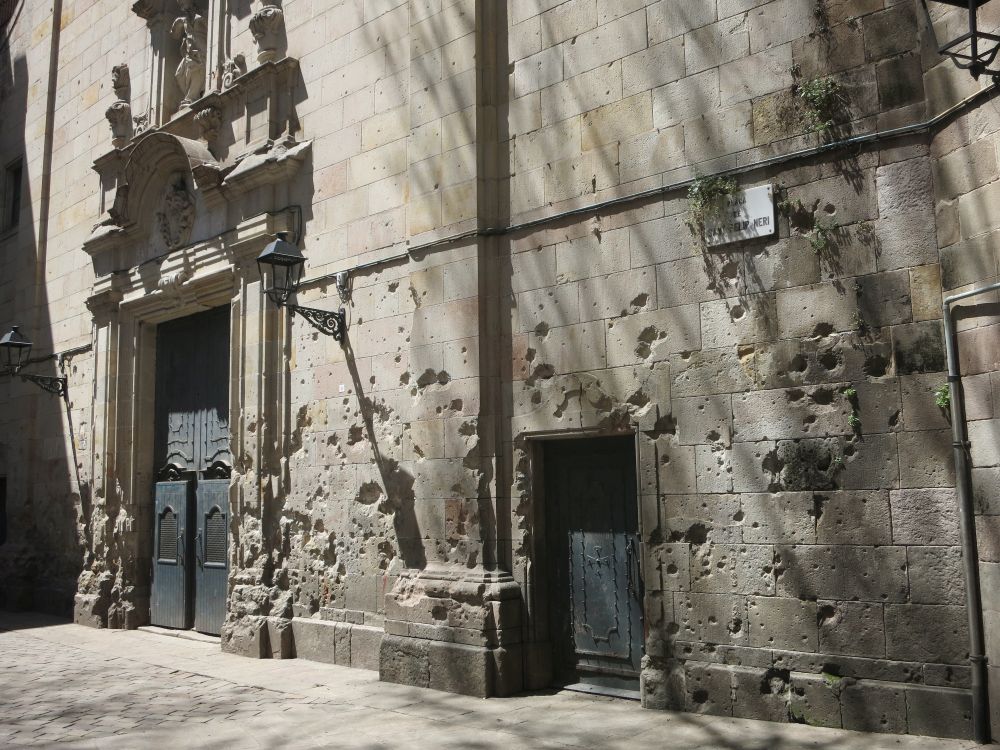 Stone wall with bullet or shrapnel holes, and an ornate doorway next to it.