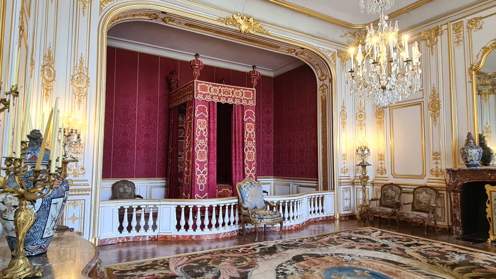 A large canopy bed in an alcove, separated from the rest of the room by a low fence. A chandelier, elegant gold-edge panelling.