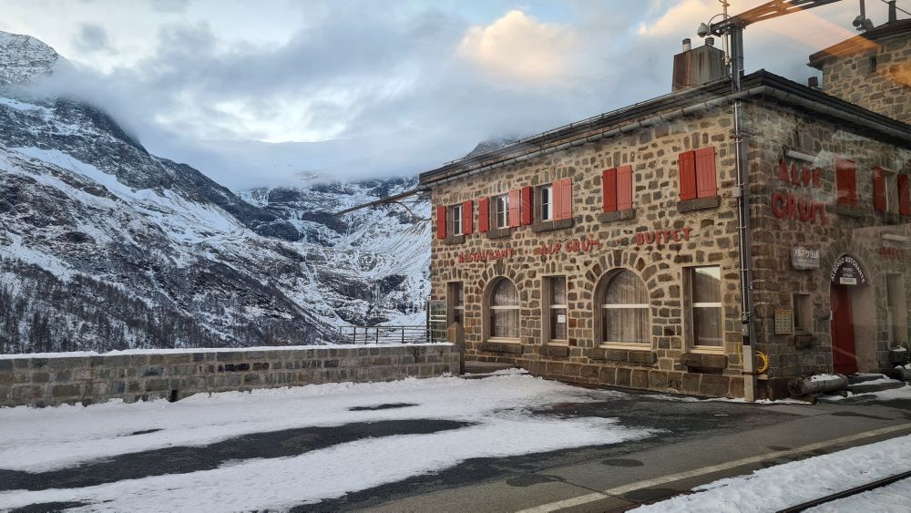 A stone building, 2 stories, and beyond it, snowy mountains, seen from the Rhaetian Railway tracks.