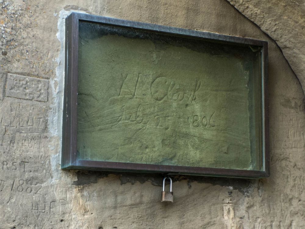 Protected by a glass frame attached to the rock, the Clark signature with other signatures carved around it.