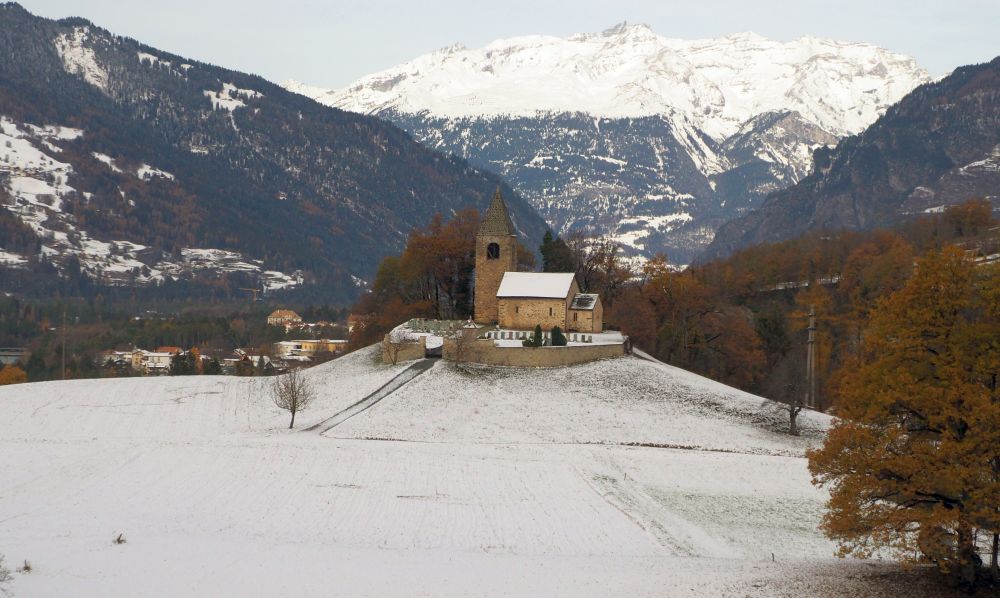 A small stone church with tower on an outcropping - snow-capped mountains behind it.