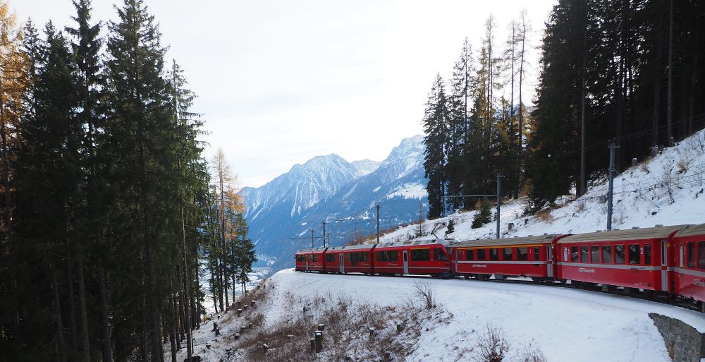 A red train curving along a track, snow-capped mountains in the distance.