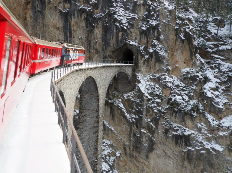 A bright red train curving atop an arched viaduct, heading for a tunnel in a wall of rock.