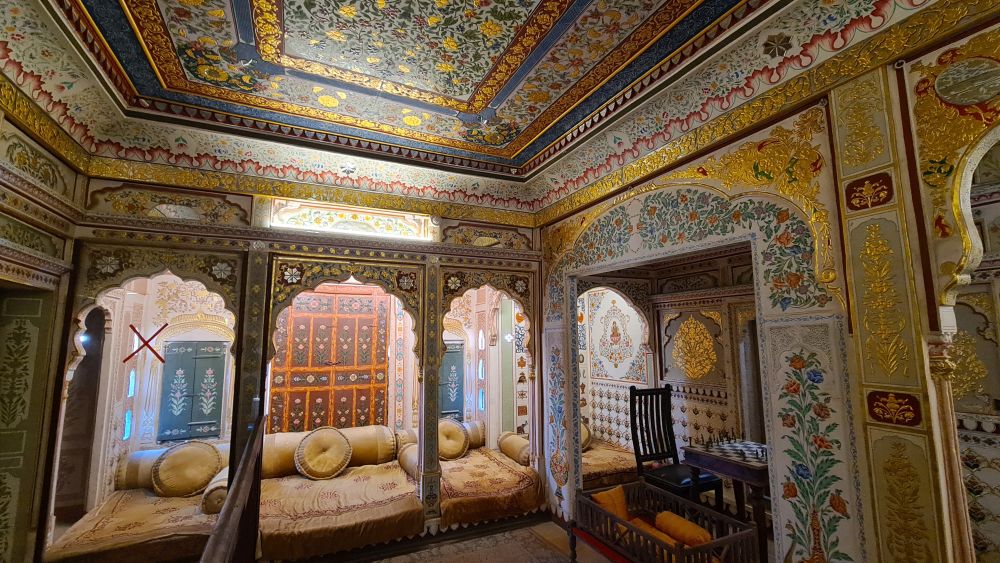 A room with arches around its sides, sofas on the floor around its sides, and detailed paint and tilework on walls and ceilings.