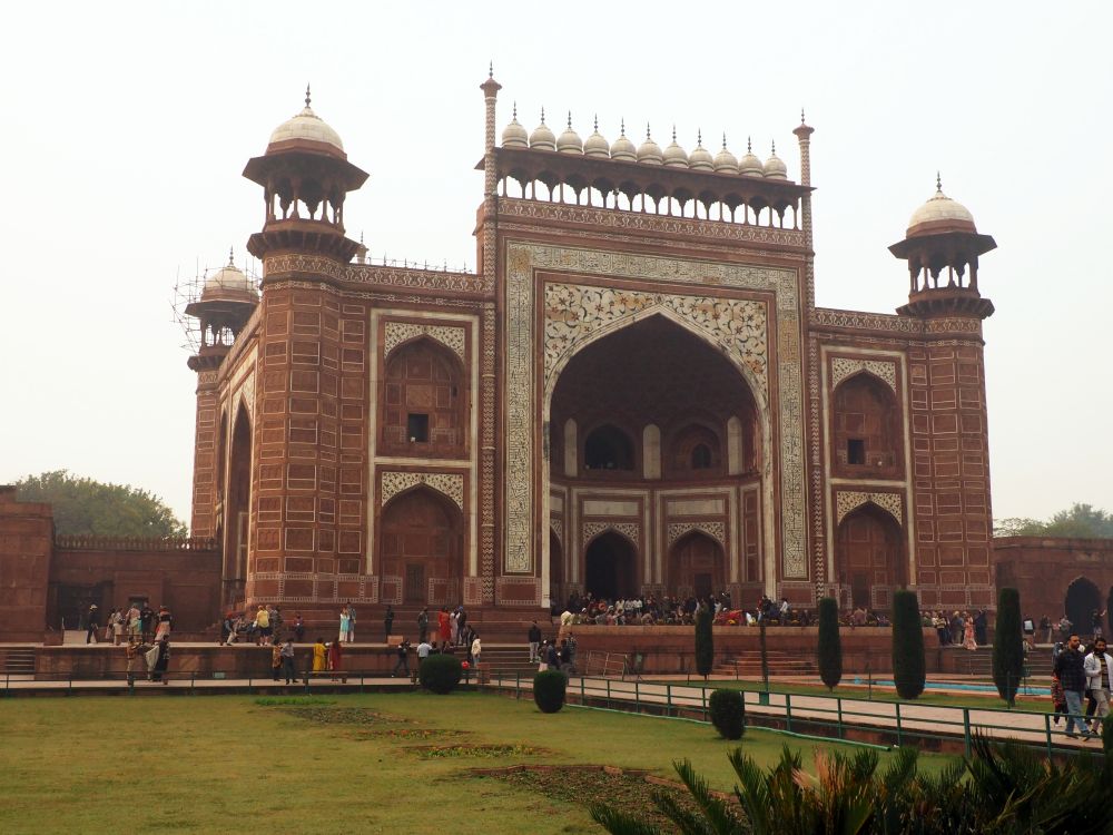 A large red sandstone building with a very high arch at its center and detailed decorations in white stone. Small domes on each corner and across the center gateway.