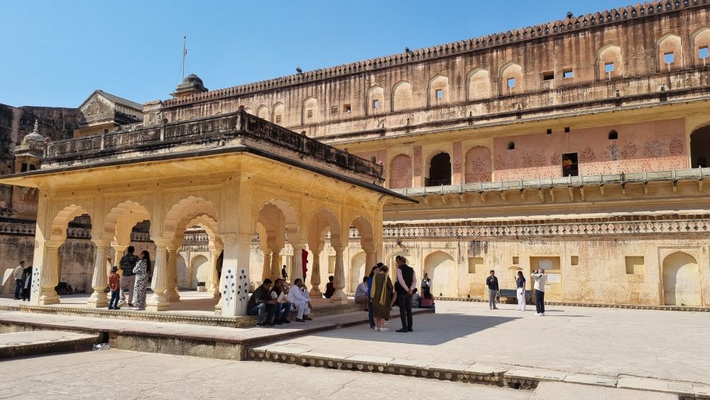 A open area with a small, arched pavilion in the center. The building behind it has several stories lined with balconies that have flower decorations on the walls.