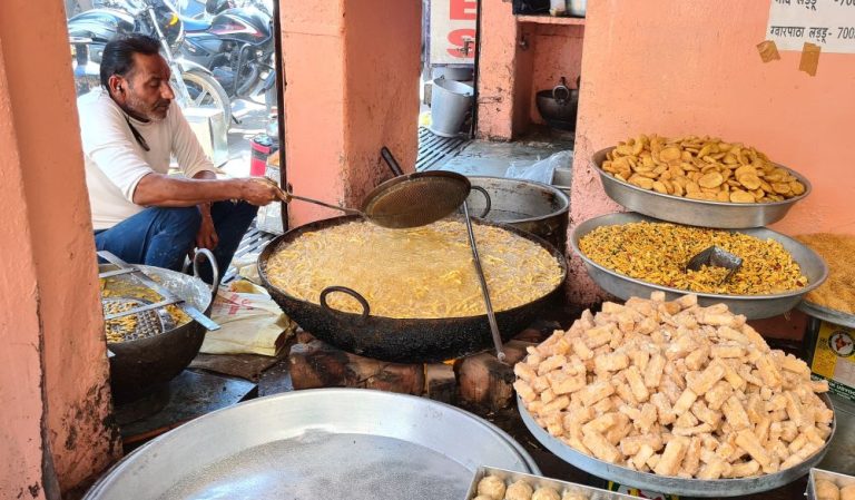 A man stirring something in a large wok, with trays of various snacks around the wok.