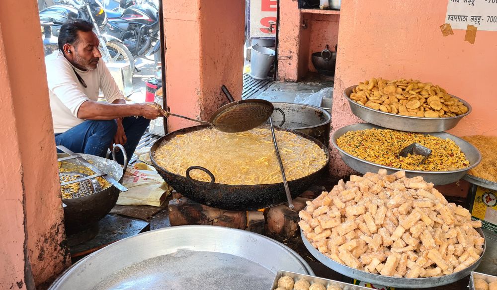 A man sits on the ground stirring something in a huge wok. Other large trays around the wok display various snacks.