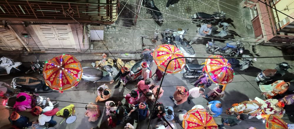 Seen from above, a group of people, colorfully dressed, some carry large umbrellas that are lit up. A drummer on the left and a man on a horse on the right.