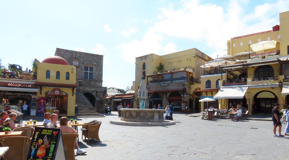 A large open plaza with a fountain in the middle and shops and cafes all around.